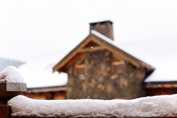 Snow-covered surface for product placement. Blurred house made of wood and stone on background....
