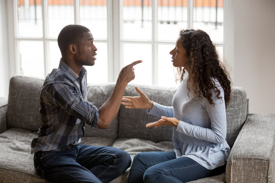 Irritated African American couple quarrelling at home, sitting together on couch, angry man blaming, negative emotionally shouting at woman making excuses, points her finger, break up, problem