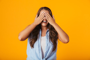Excited young woman posing isolated over yellow wall background covering eyes with hands.