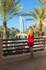 Rolgordijnen Young tourist woman dressed in red dress looking Burj al Arab from Madinat Jumeirah souk with shopping bags next to her. Beautiful destination in Dubai. © DanRentea