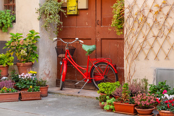 Fototapeta na wymiar Red bicycle near picturesque door in the street. Old street of medieval town Pienza are decorated with flowers and ivy, Italy