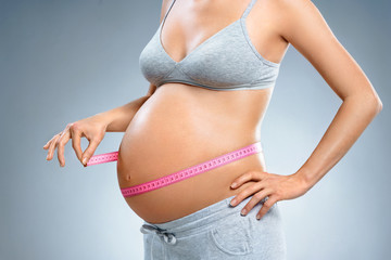 Close up of pregnant woman with measuring tape measures her belly on grey background. Pregnancy, maternity, preparation and expectation concept