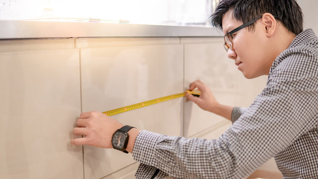 Young Asian man using tape measure for measuring modern kitchen counter in showroom. Shopping furniture for home improvement concept