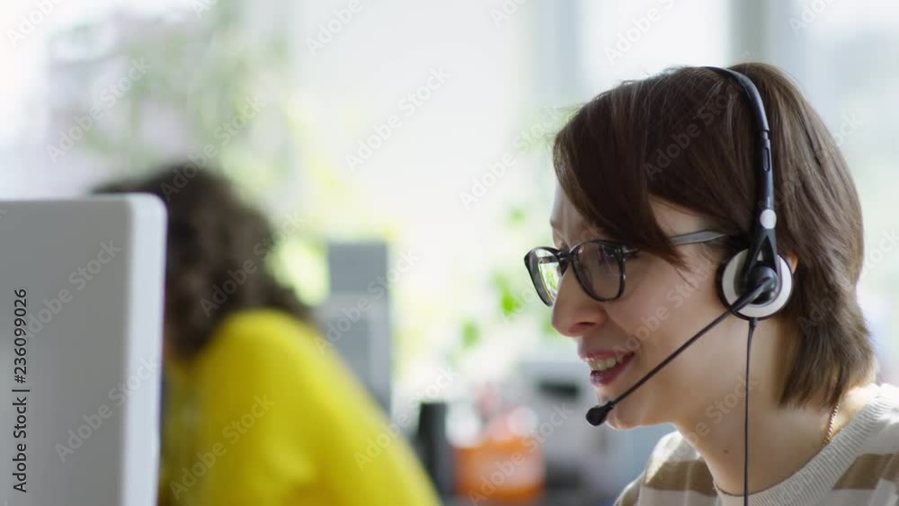 Poster tilt down of young caucasian woman in headset with mic using computer and talking with client while 