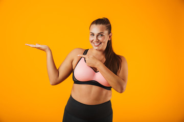 Portrait of european chubby woman in sportive bra holding copyspace on palm, isolated over yellow...