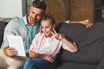 father holding digital tablet while daughter showing peace sign during video call