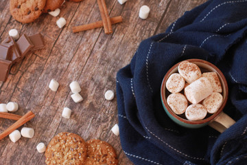 Hot drink with marshmallow wrapped in scarf. Autumn and winter drink concept. Cookies, cinnamon and pieces of chocolate scattered on vintage wooden background.