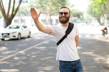 Joyful hipster guy gesturing good bye. Bearded young man in sunglasses and summer shirt standing on road and raising hand. Gesturing concept