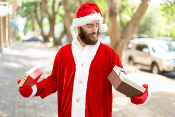 Pensive Christmas courier guessing what in gift boxes. Bearded young man wearing Santa costume carrying presents and staring at boxes. Christmas gifts concept