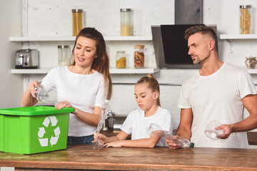 Obraz premium family standing at kitchen and putting empty plastic bottles in recycle box