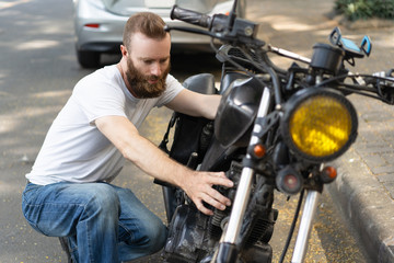 Obraz premium Serious biker diagnosing broken motorbike. Bearded young man sitting on haunches and examining motorcycle. Breakdown concept