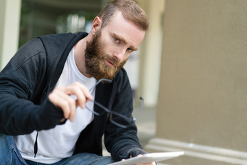 Serious pensive hipster guy resting during walk. Bearded young man putting on his glasses and using tablet outdoors. Communication concept