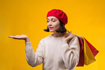 Shopping woman showing something with open hand palm. Happy girl holding shopping bags. Fashion woman shopper isolated on yellow background. Pretty woman in red beret. Copy space. Seasonal sales.