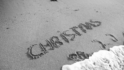Black and white photo of word Christmas written on wet san on the beach. Sea wave flowing over. Concept of winter holidays, New Year and tourism
