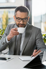 businessman in suit and eyeglasses drinking coffee and writing in notebook at wokrplace