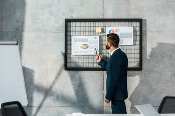 side view of bearded businessman holding pen and pointing at business charts on wall