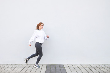 Happy girl wearing sportswear running against the background of a white wall. Young attractive woman is running on the street on a white background. Copyspace