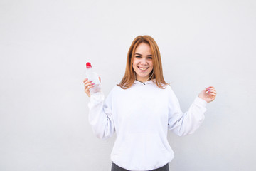 Portrait of a happy, attractive girl in sportswear and a bottle of water in her hands, listens to music in the headphones, looks at the camera and smiles. Joyful sports girl on a white background