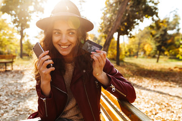 Beautiful cute woman sitting on a bench in park using mobile phone holding credit card.