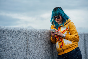 Portrait of young woman with dyed blue hair leaning against wall looking at cell phone