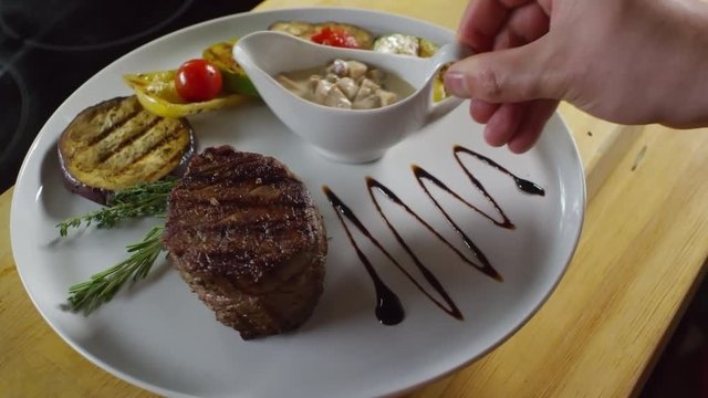 Handheld close up shot of unrecognizable cook putting sauce dish on plate with fillet mignon and vegetables