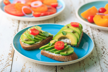 Avocado sandwich on dark rye toast bread made with fresh sliced avocado, cream cheese, seeds and cherry on white wooden background