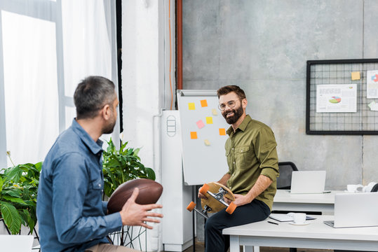 smiling businessmen looking at each other and holding american football ball and longboard in office