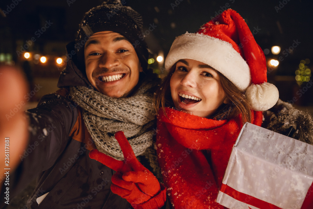 Wall mural young loving couple sitting outdoors in evening in christmas hat holding gift box take a selfie by c