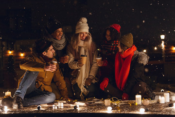 Happy young group of friends sitting outdoors in evening in christmas hats drinking coffee.