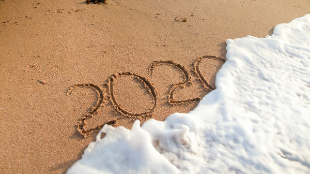 Closeup photo of sea waves rolling over 2020 New Year numbers written on wet sand