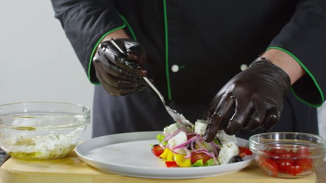 Mid-section shot of hands of unrecognizable male chef in uniform and gloves putting cubes of marinated feta cheese on salad made of fresh vegetables