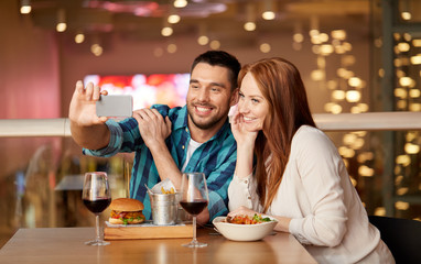 leisure, technology and people concept - happy couple having lunch and taking selfie by smartphone at restaurant