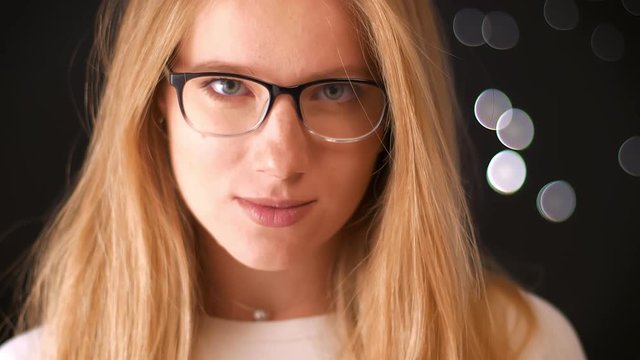 Charming flirting caucasian blonde woman is standing straight and confidently looking at camera above her glasses while hanging in black studio, focused