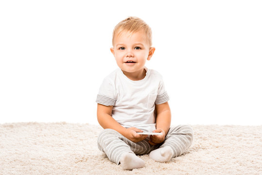 happy toddler boy sitting on carpet and holding smartphone isolated on white