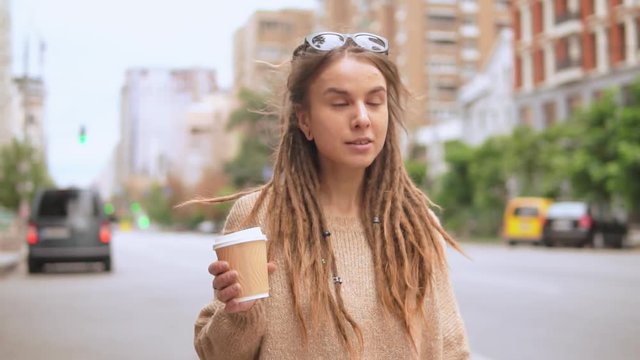 hipster girl holding cup of coffee to go walking on the street .smiling caucasian model with dreadlocks wearing oversized sweater going along the road in the morning