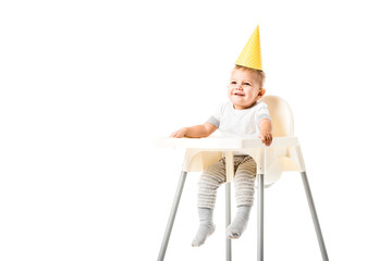happy toddler boy with yellow party hat on head sitting in highchair and smiling isolated on white