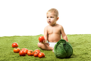 Fototapeta premium adorable toddler boy holding tomato and sitting on green carpet with vegetables isolated on white