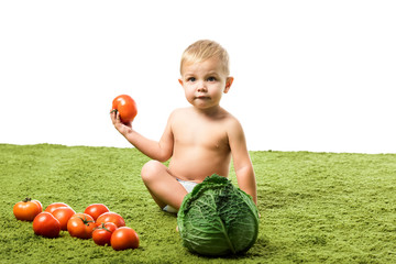 Fototapeta premium toddler boy holding tomato and sitting on green carpet near vegetables isolated on white