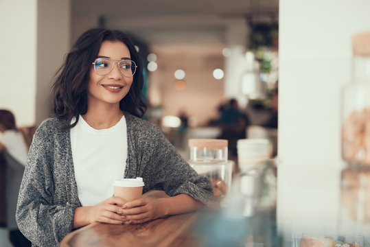 Cute young lady holding cup of coffee and smiling while sitting alone
