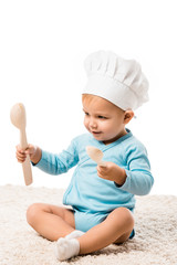 toddler boy in chefs hat sitting on carpet, holding two big wooden spoons and smiling isolated on white
