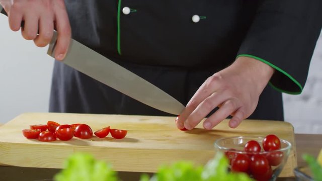 Mid-section tracking shot of unrecognizable cook in uniform cutting cherry tomatoes with sharp knife on wooden board