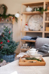Cozy home interior and decoration, tea time. Wooden serving tray on sofa near Christmas tree with red ornaments. Morning breakfast with mug of hot beverages, white teapot in living room.