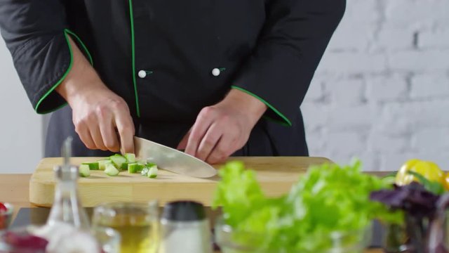 Mid-section PAN shot of unrecognizable male restaurant chef in uniform chopping cucumber on wooden board