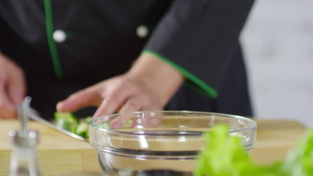 Close up shot of hands of unrecognizable male cook in uniform putting chopped cucumber into glass bowl while preparing salad