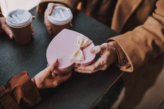 Close up of heart-shaped gift box with bow in hands of senior couple. Pensioners sitting at the table with cups of coffee