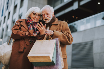 Portrait of stylish lady holding smartphone while husband with shopping bags pointing at display....