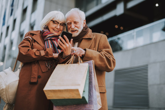 Portrait of stylish lady holding smartphone while husband with shopping bags pointing at display. They watching videos and smiling