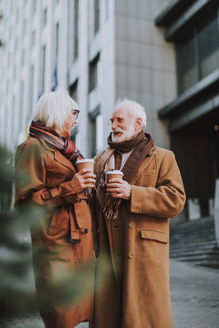Portrait of stylish old gentleman spending time with wife outdoors. They holding cups of coffee while looking at each other and smiling