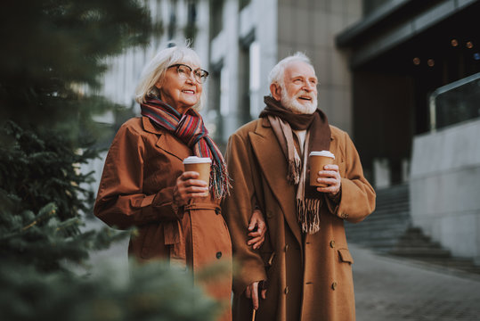 Portrait of stylish old lady in glasses spending time with husband outdoors. They holding cups of coffee while looking away and smiling
