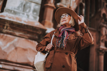 Obraz premium Street style. Low angle portrait of lovely senior lady with backpack standing on the street and touching hat. She is looking away and smiling